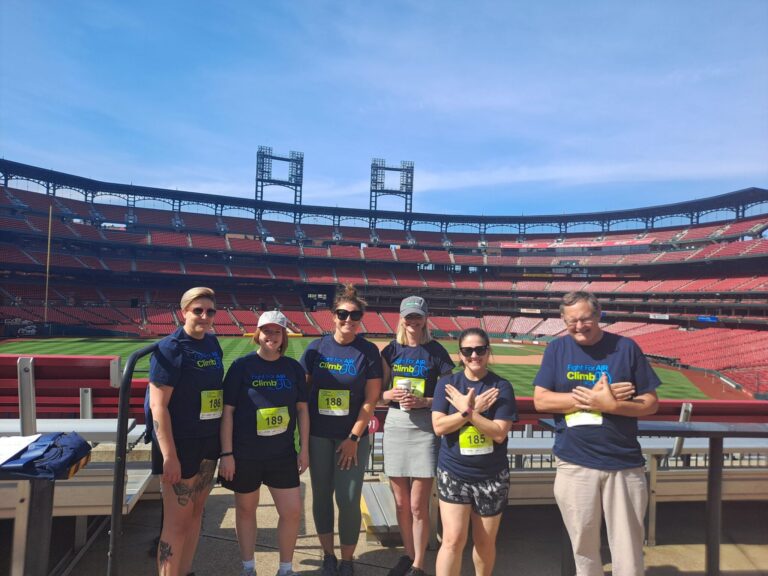 Six people wearing matching navy blue "Fight For Air Climb 30" t-shirts and yellow race bibs stand in front of a baseball field inside a large stadium with red seats. The group is posing with some crossing their arms in front of their chests. The sky is clear and blue.
