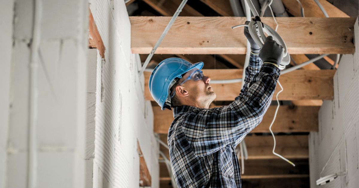 A construction worker wearing a blue hard hat, safety glasses, gloves, and a plaid shirt is installing or inspecting electrical wiring in a building with exposed wooden beams and white brick walls.