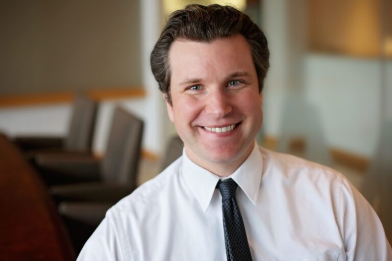 Smiling man with neatly styled dark hair wearing a white dress shirt and a dark textured tie, seated in a softly lit office or conference room with blurred chairs and walls in the background.