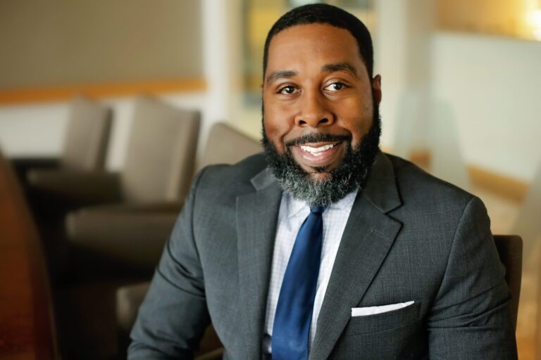 Man with a beard wearing a dark gray suit, white checkered shirt, and navy blue tie, smiling while seated in an office setting with blurred chairs in the background.