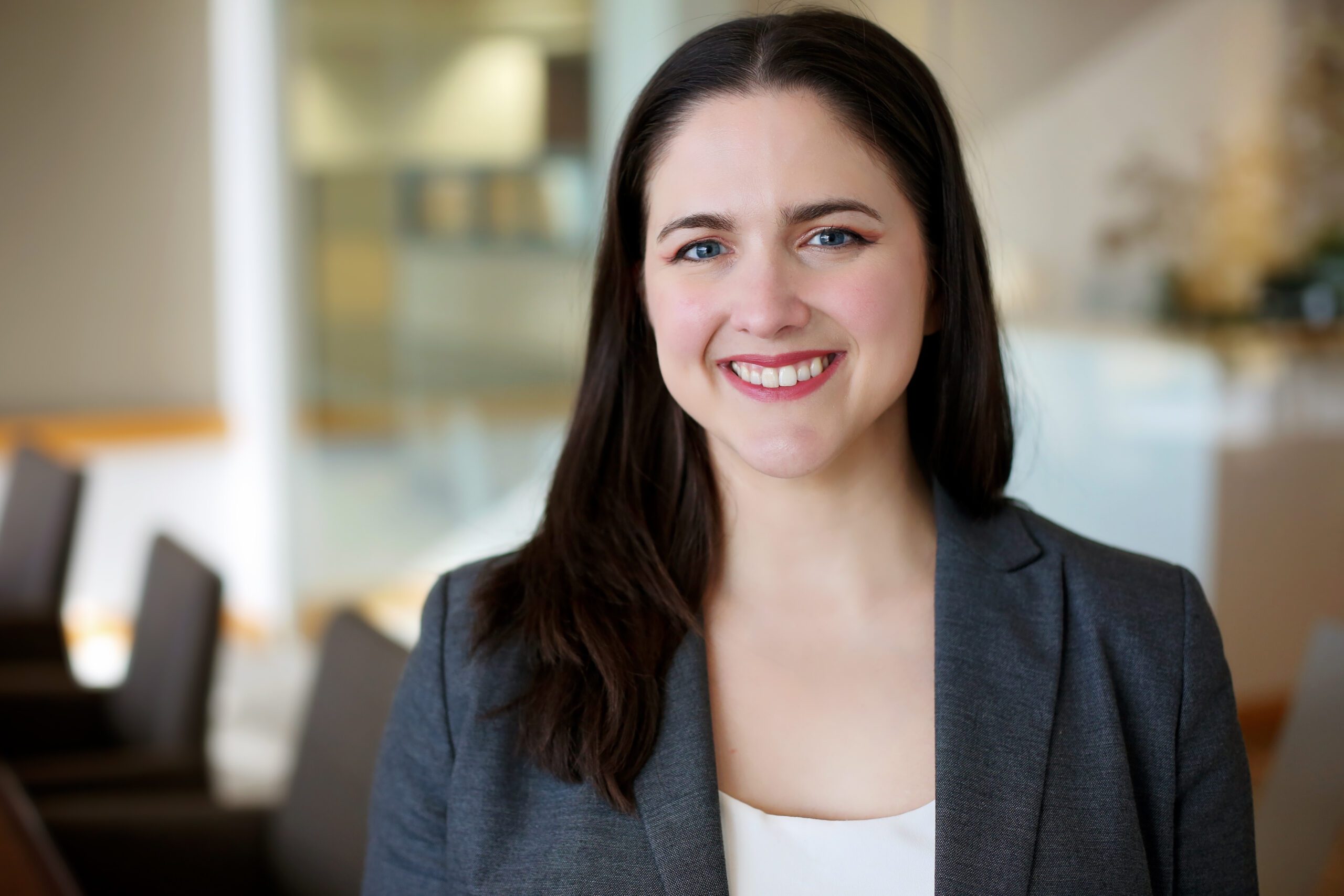 A smiling woman with long dark hair wearing a gray blazer over a white top, standing in a softly blurred indoor setting with chairs and light-colored walls in the background.