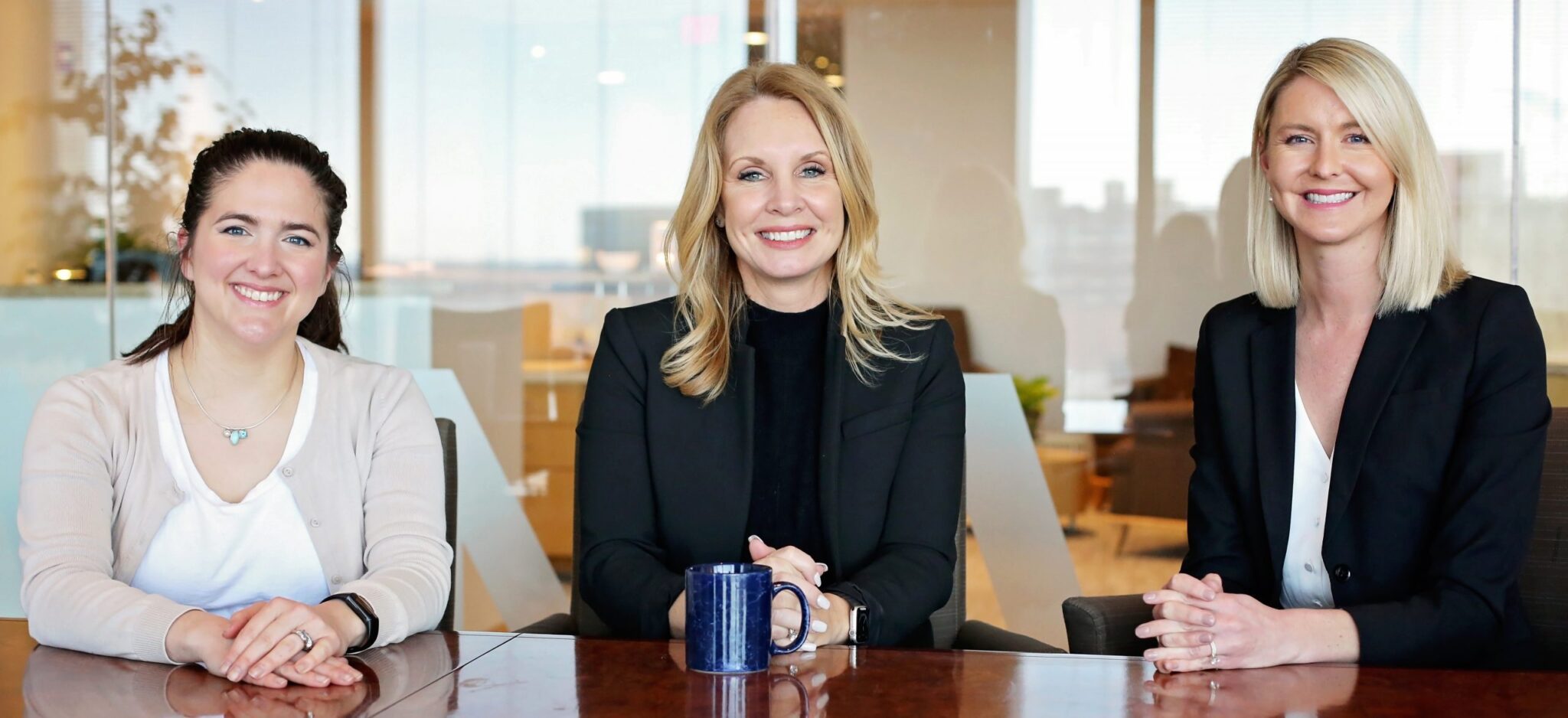 Three women sitting at a wooden conference table in an office setting. The woman on the left has dark hair tied back, wearing a light beige cardigan over a white top, and a necklace. The woman in the center has blonde hair, wearing a black blazer and black top, with a blue mug in front of her. The woman on the right has straight blonde hair, wearing a black blazer over a white blouse. All three are smiling and have their hands resting on the table.