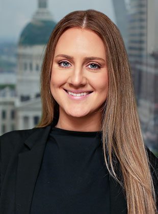 Young woman with long straight light brown hair, wearing a black top and black blazer, smiling in front of a blurred cityscape background.