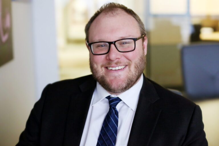 Smiling man with light skin, short light brown hair, and a beard, wearing black rectangular glasses, a black suit jacket, a white dress shirt, and a dark blue striped tie, seated in an office setting with blurred background.