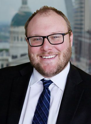 Man with light skin, short light brown hair, and a beard, wearing black rectangular glasses, a black suit jacket, a white dress shirt, and a dark blue striped tie, smiling in front of a blurred cityscape background.
