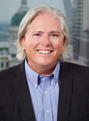 A middle-aged person with shoulder-length gray hair, smiling. They are wearing a light blue button-up shirt under a dark checkered blazer. The background shows an out-of-focus cityscape with buildings and a river.