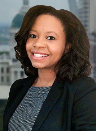 A woman with shoulder-length curly dark hair is smiling. She is wearing a black blazer over a patterned top. The background shows a blurred cityscape with buildings and a dome structure.