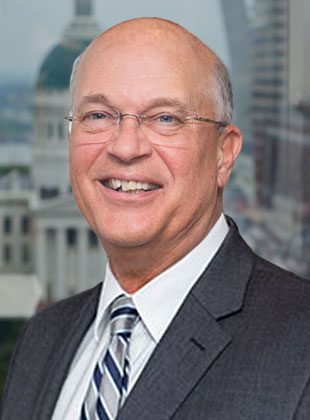 Middle-aged man wearing glasses, a dark suit, white shirt, and striped tie, smiling with a cityscape background featuring a domed building.