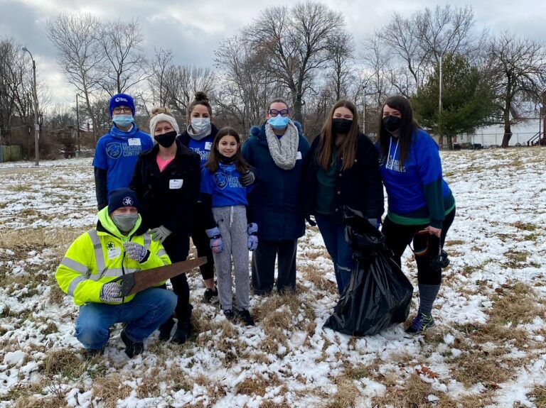 A group of eight people standing outdoors on a snowy grassy field, dressed warmly in winter clothing and face masks. Most are wearing blue shirts with "The Mission Continues" logo. One person in a bright yellow jacket is crouching in front holding a saw, while another person holds a large black trash bag. Leafless trees and a cloudy sky are visible in the background.