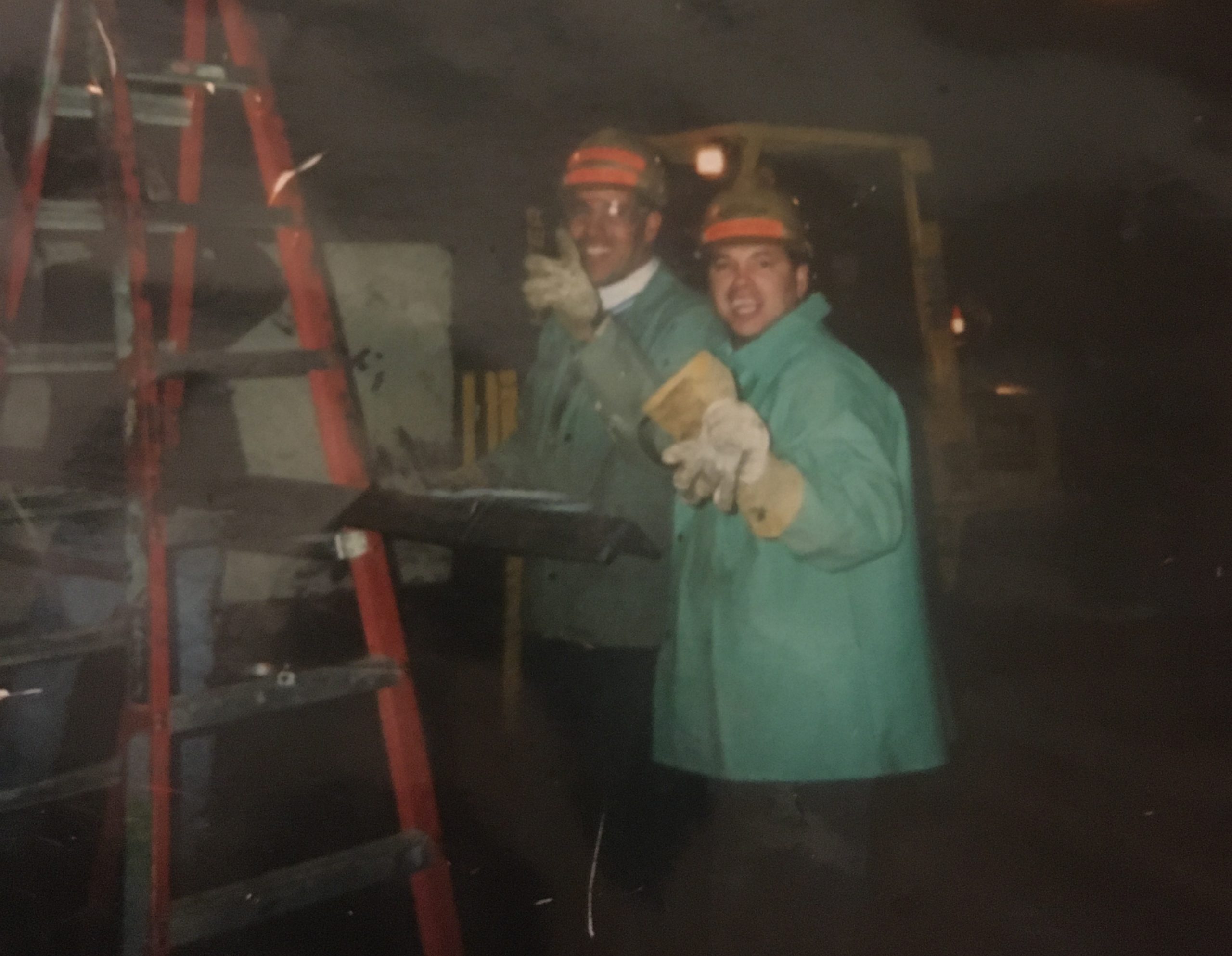 Two workers wearing green protective jackets, gloves, and helmets with orange stripes are standing next to a red ladder in a dimly lit industrial or construction setting. One worker is holding a tool or object, and both appear to be engaged in their task.
