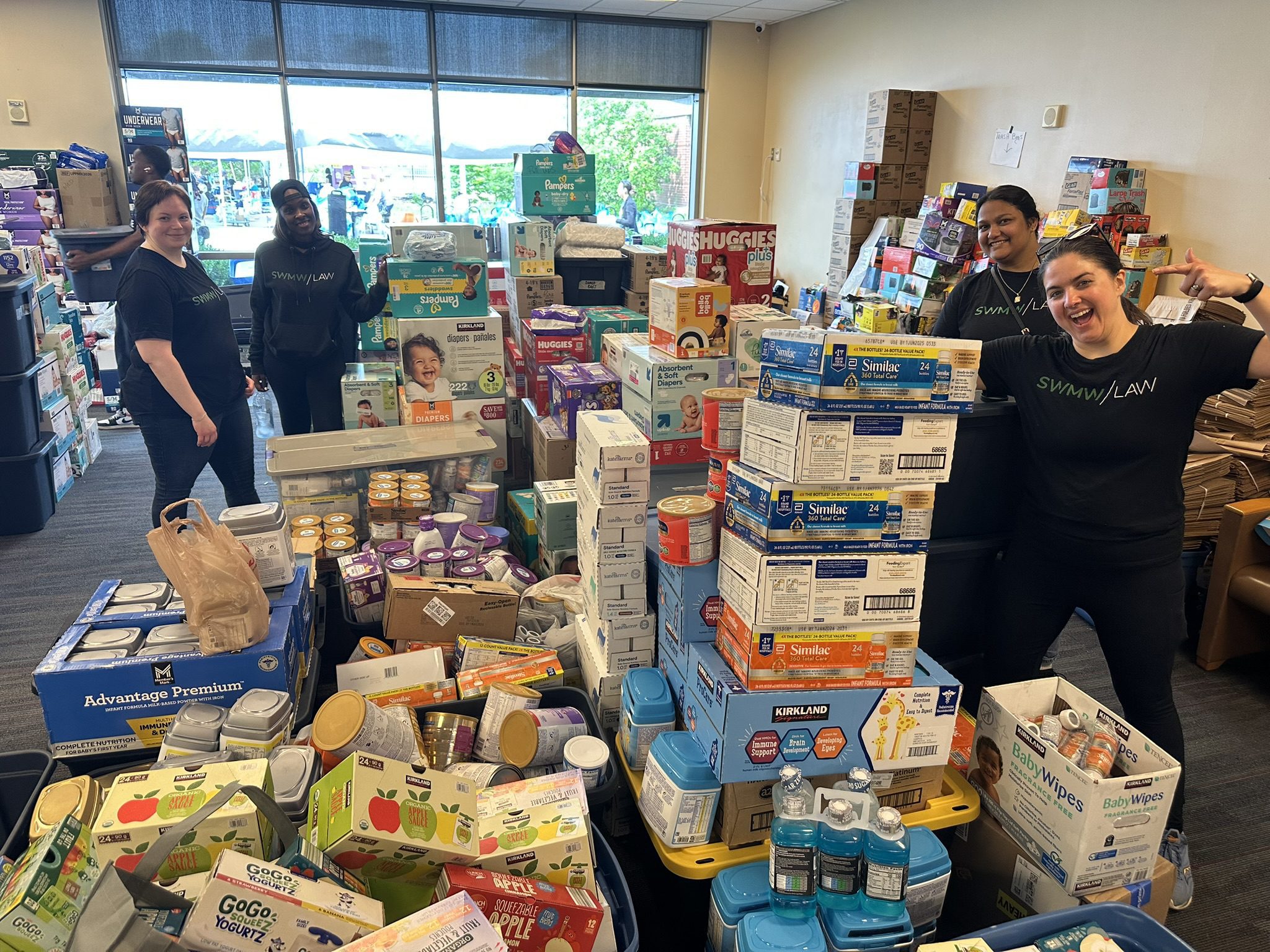 Four people wearing black "SWMW/LAW" shirts stand among large stacks of baby supplies, including diapers, baby formula, baby wipes, and snacks, organized in a room with large windows. The supplies are arranged in boxes and containers, filling much of the floor space.