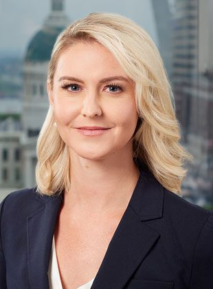 Blonde woman with shoulder-length wavy hair, wearing a dark blazer over a white top, smiling slightly, with a blurred cityscape background featuring a domed building.