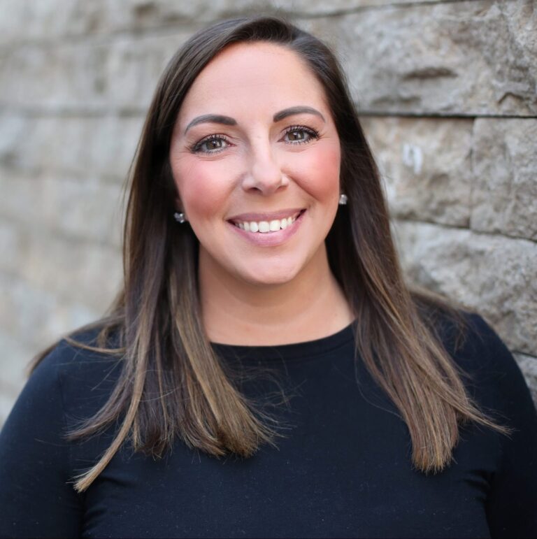 Woman with straight, shoulder-length brown hair wearing a black top, smiling, and standing in front of a textured stone wall.