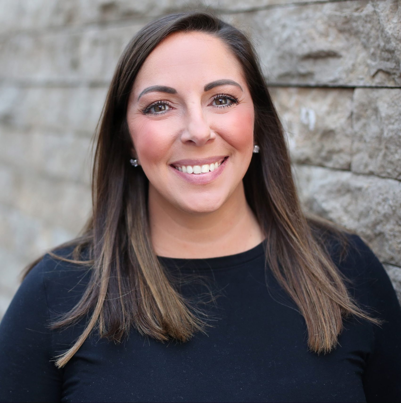Woman with straight, shoulder-length brown hair wearing a black top, smiling, and standing in front of a textured stone wall.