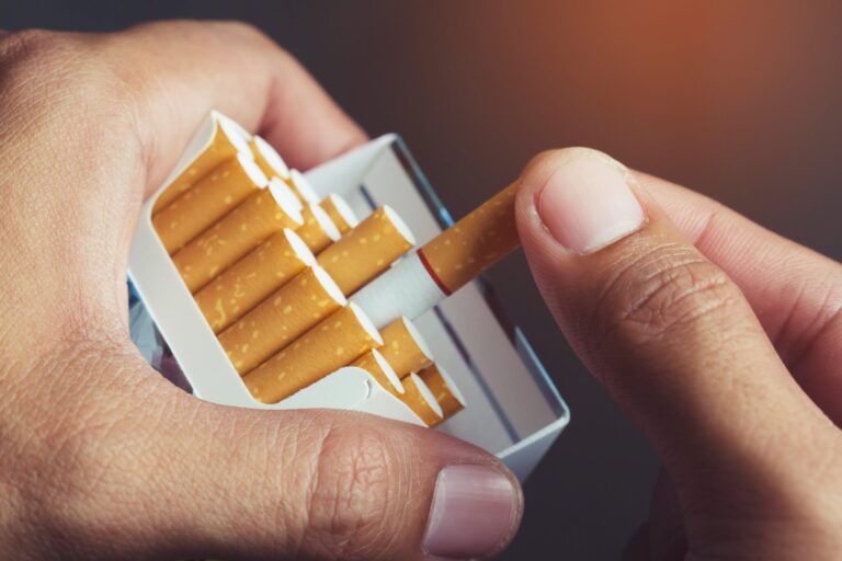 A close-up of a hand holding an open pack of cigarettes, with the thumb and index finger of the other hand pulling out a single cigarette. The cigarettes have brown filters and white paper. The background is blurred with a warm tone.