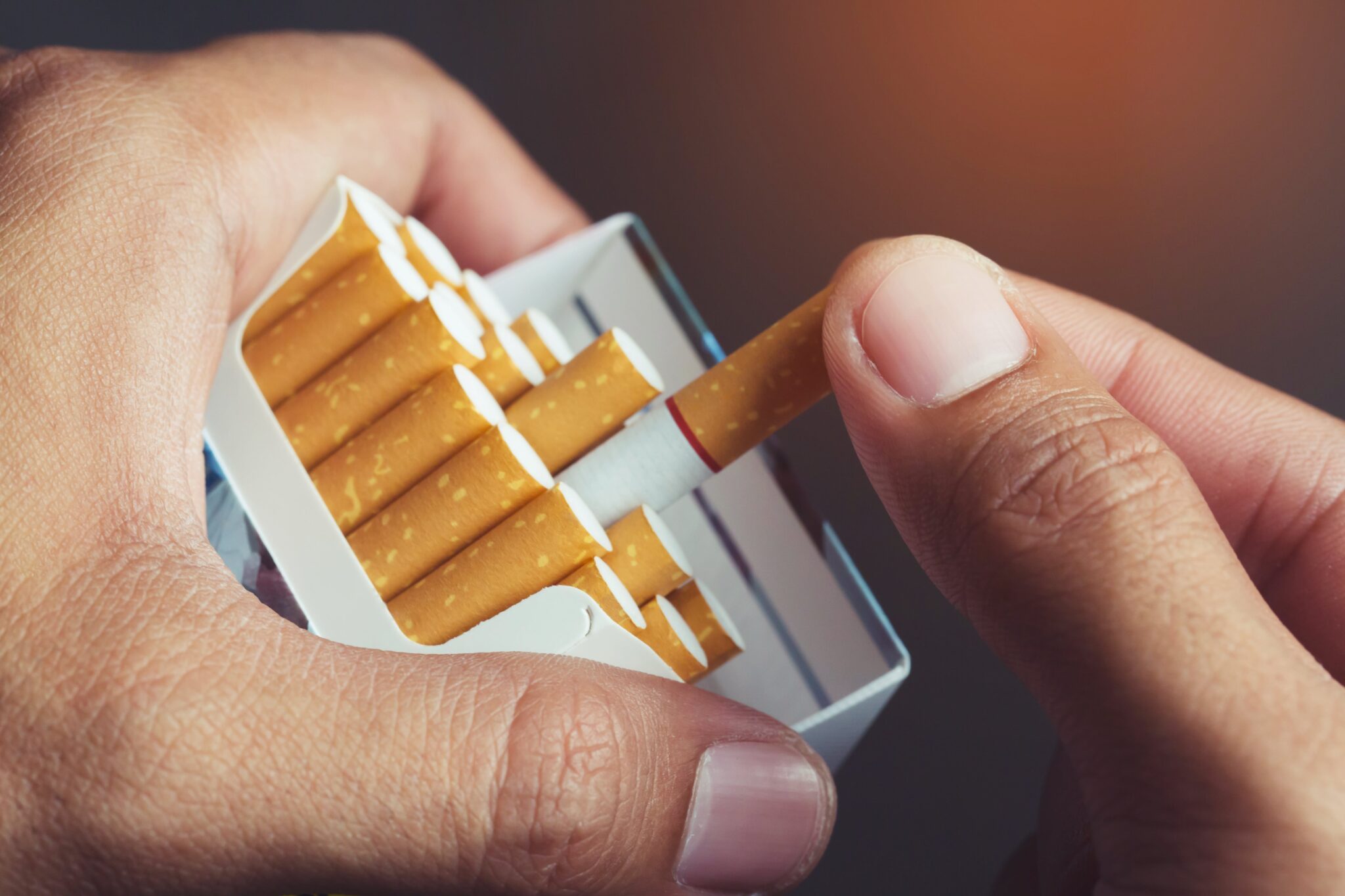 A close-up of a hand holding an open pack of cigarettes, with the thumb and index finger of the other hand pulling out a single cigarette. The cigarettes have brown filters and white paper. The background is blurred with a warm tone.