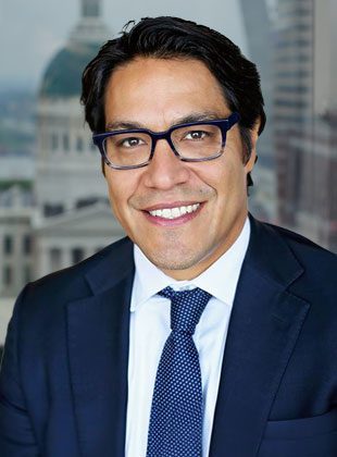 Man with dark hair and glasses wearing a dark blue suit, white shirt, and blue patterned tie, smiling in front of a blurred cityscape background.