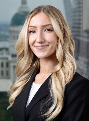 Young woman with long, wavy blonde hair wearing a black blazer and white top, smiling softly. The background shows an out-of-focus cityscape with buildings.