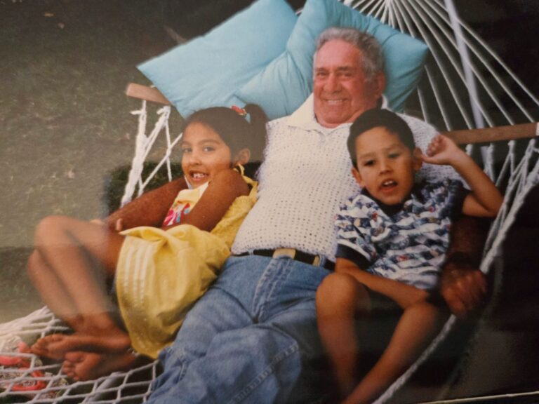 An older man with gray hair wearing a white knit shirt and blue jeans is lying on a hammock with two children. The girl on the left is wearing a yellow dress and has her hair tied back, while the boy on the right is wearing a patterned shirt and shorts. They are all smiling and appear relaxed, with light blue pillows behind the man's head.