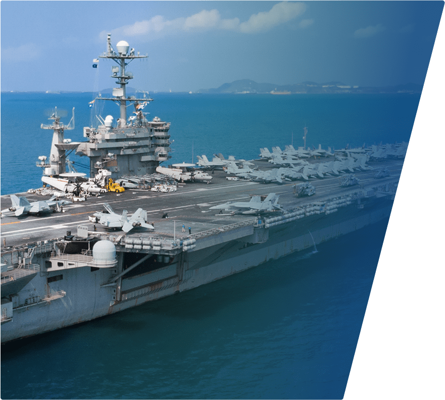 Aircraft carrier at sea with multiple fighter jets parked on the flight deck, a control tower, and various equipment visible. The background shows calm blue water and distant land under a partly cloudy sky.