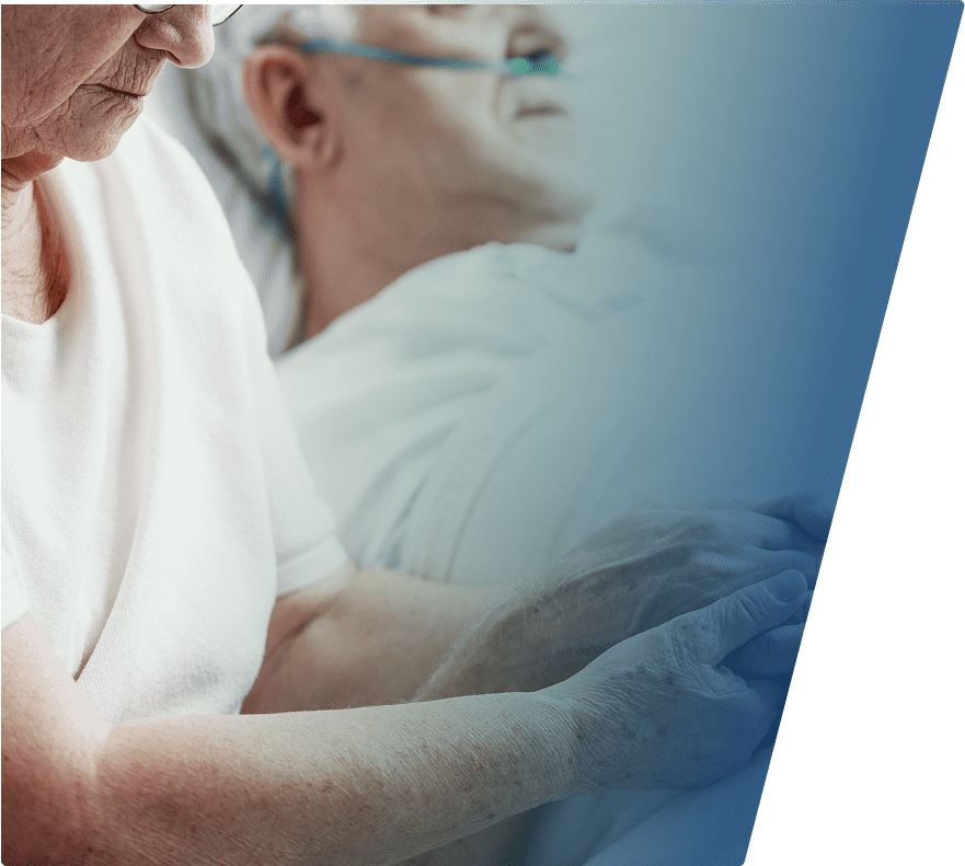 Two elderly individuals are shown close-up, with one person gently holding the other's hand. Both are wearing light-colored clothing, and the scene conveys a sense of care and support.