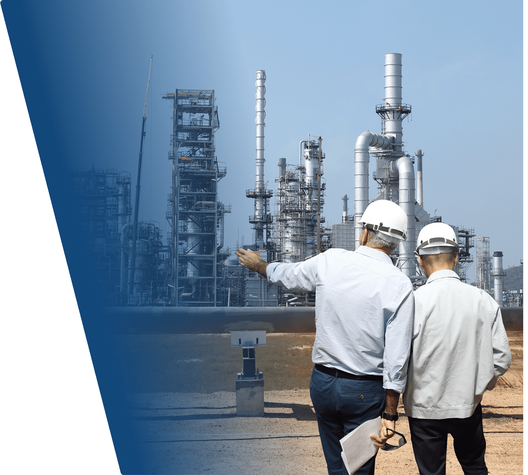 Two engineers wearing white hard hats and light-colored shirts are standing outdoors at an industrial site, facing a large complex of metal structures and tall chimneys. One engineer is pointing towards the facility while holding documents and safety glasses in the other hand. The background shows a clear sky and the industrial equipment appears to be part of a refinery or chemical plant.