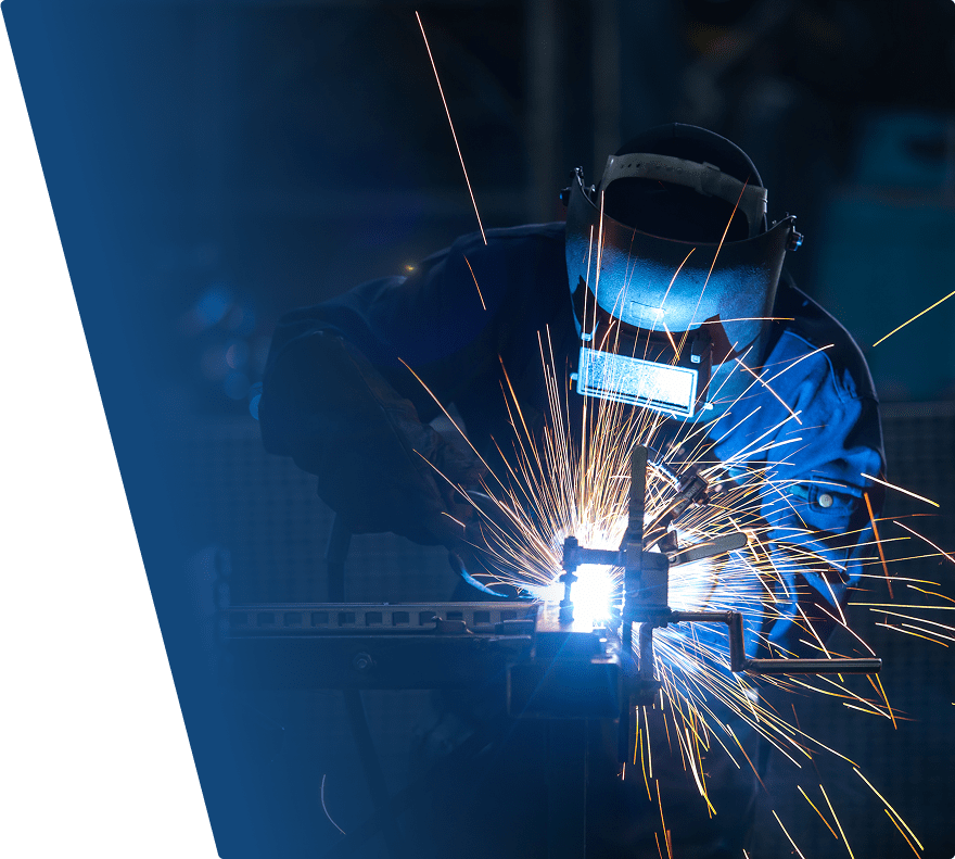 A welder wearing protective gear, including a welding helmet and gloves, is actively welding metal, producing bright sparks and intense blue light. The background is dark, emphasizing the vivid sparks and the focused work.
