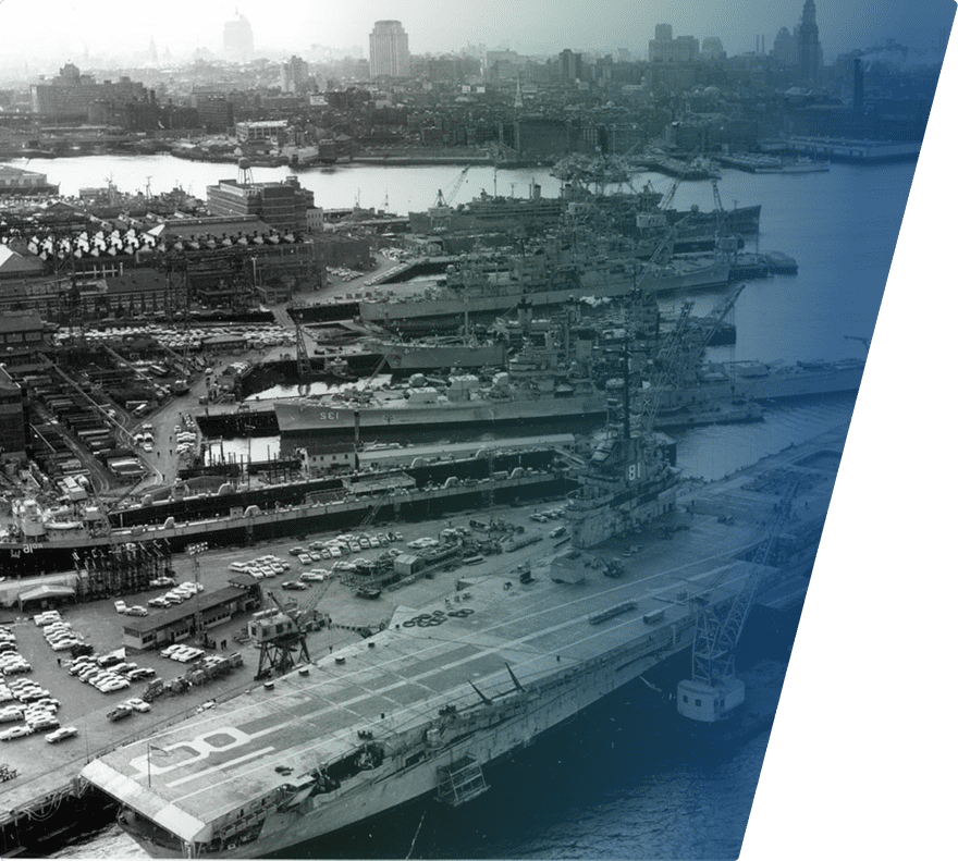 A large naval shipyard with multiple warships docked, including a prominent aircraft carrier in the foreground marked with the number "81." The area is busy with numerous vehicles and cranes, and a city skyline is visible in the background under a hazy sky.