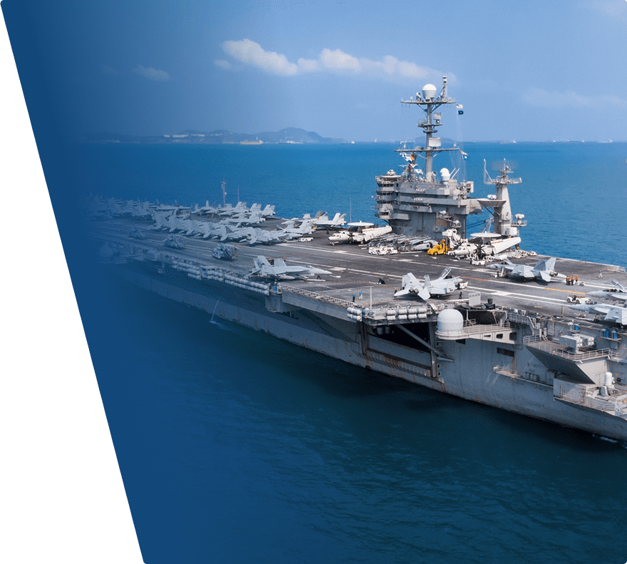 A large aircraft carrier sailing on the ocean with multiple fighter jets parked on its deck. The ship has a tall superstructure with radar and communication equipment, and the sea is calm with a distant coastline visible under a partly cloudy sky.