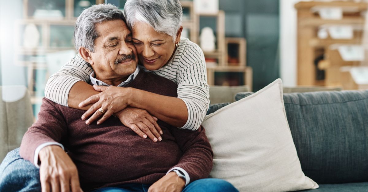 An elderly couple sitting on a couch, with the woman hugging the man from behind. Both have gray hair, the man is wearing a maroon sweater and blue jeans, and the woman is wearing a white and black striped top. They appear content and affectionate, with a beige pillow on the couch beside them. The background shows a cozy, softly lit living room.