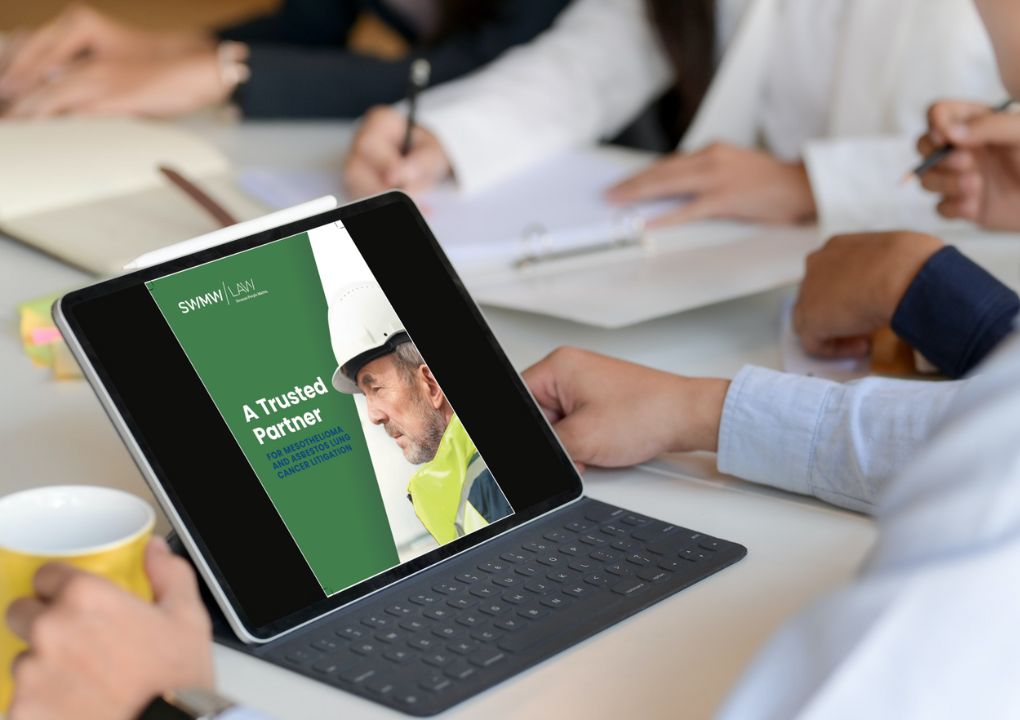 A tablet with a keyboard case displaying a digital brochure titled "A Trusted Partner for Mesothelioma and Asbestos Lung Cancer Litigation" featuring a man in a white hard hat and yellow safety vest. The tablet is on a table surrounded by people writing on paper and holding a yellow cup.