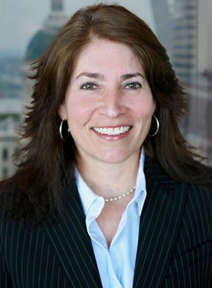 Smiling woman with medium-length brown hair wearing a black pinstripe blazer, light blue collared shirt, pearl necklace, and hoop earrings, with a blurred warm-toned background.