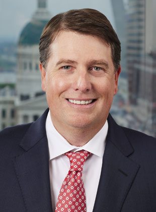 Man with short brown hair wearing a dark navy suit, white dress shirt, and red patterned tie, smiling in front of a blurred cityscape background featuring a domed building.