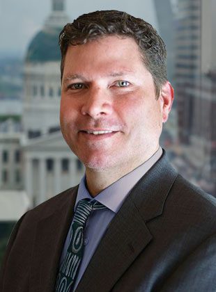 Man with short curly brown hair wearing a dark suit jacket, light purple shirt, and patterned tie, standing in front of a window with a blurred cityscape background featuring a domed building.