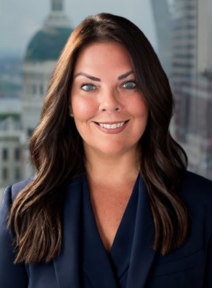 A woman with long, dark brown hair styled in loose waves, wearing a dark navy blazer. She has light skin, blue eyes, and is smiling. The background shows a blurred cityscape with a domed building and other architectural structures.