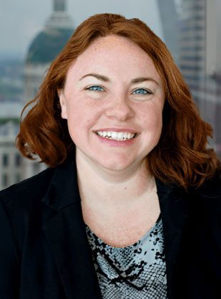 Smiling woman with shoulder-length red hair and blue eyes, wearing a black blazer over a patterned top, with a blurred cityscape background.