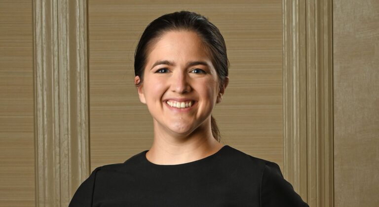 A woman with dark hair pulled back is smiling. She is wearing a black top and standing in front of a beige textured wall with vertical decorative panels.