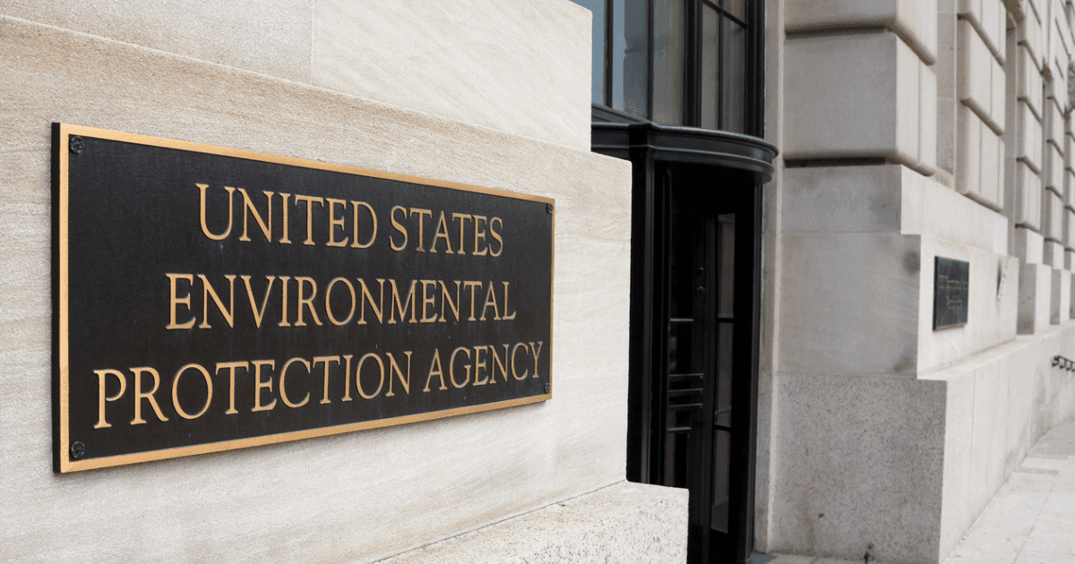 A black and gold plaque mounted on a light stone wall reads "UNITED STATES ENVIRONMENTAL PROTECTION AGENCY." The plaque is near a building entrance with large windows and a revolving door. The building exterior is made of light-colored stone blocks.
