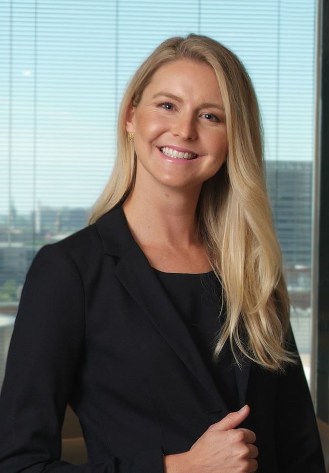 Blonde woman with long hair wearing a black blazer and black top, smiling in front of a window with horizontal blinds and a cityscape background.