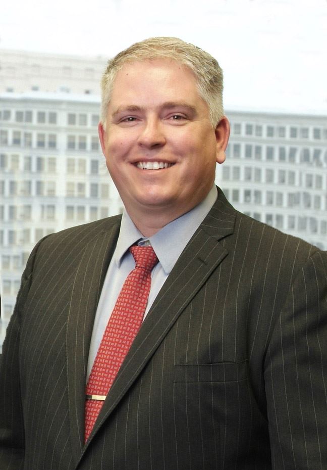 Man with short gray hair wearing a dark pinstripe suit, light blue dress shirt, and red patterned tie, smiling in front of a blurred office building background.