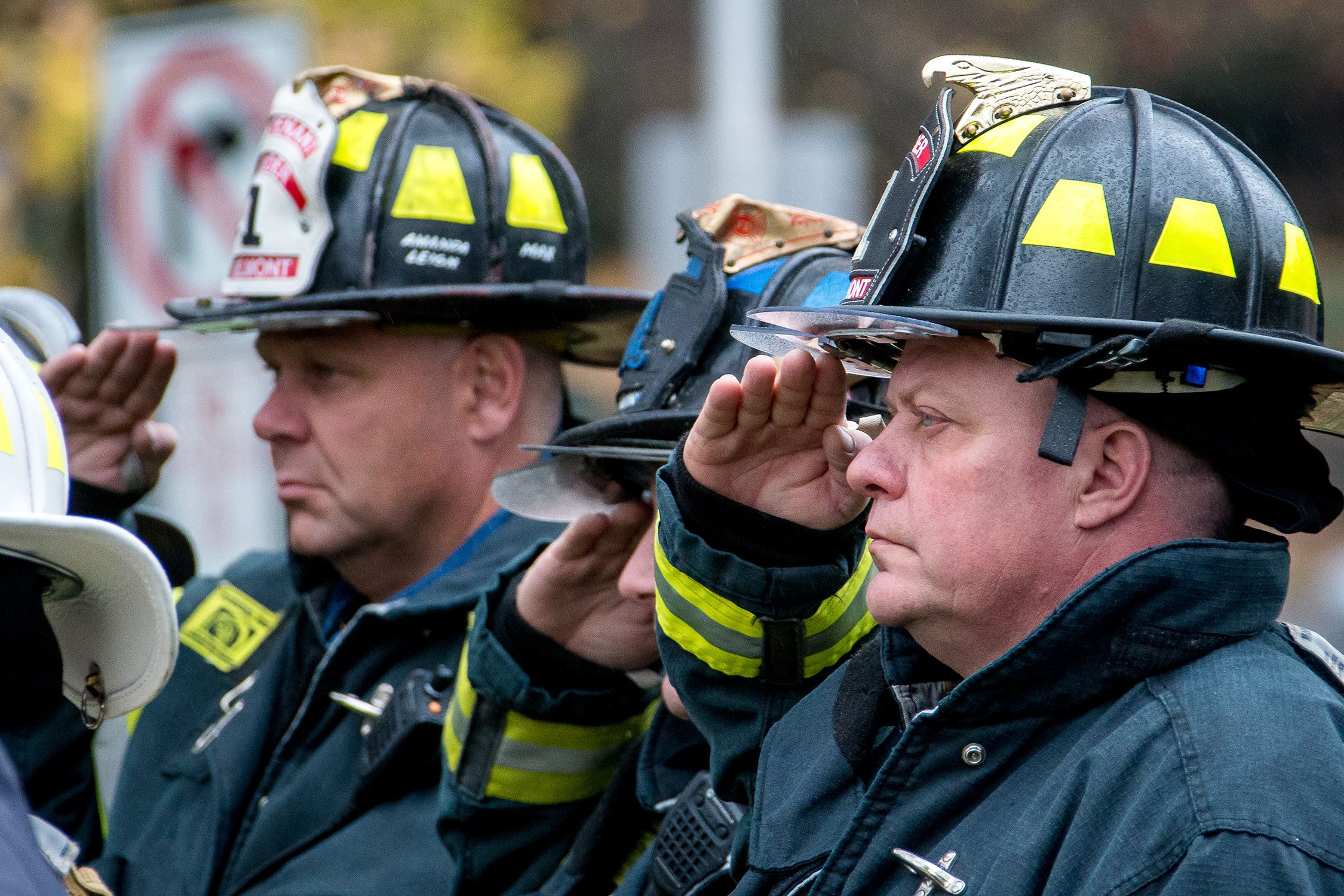 Three firefighters in uniform and helmets are standing in a row, saluting. The focus is on the firefighter closest to the camera, who has a serious expression. The helmets are black with yellow reflective markings and badges on the front. The background is blurred.