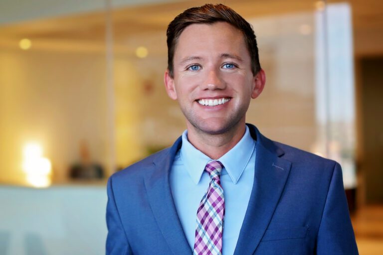 A young man with short brown hair and blue eyes is smiling. He is wearing a blue suit jacket, a light blue dress shirt, and a purple and white checkered tie. The background is softly blurred, suggesting an indoor setting with warm lighting.
