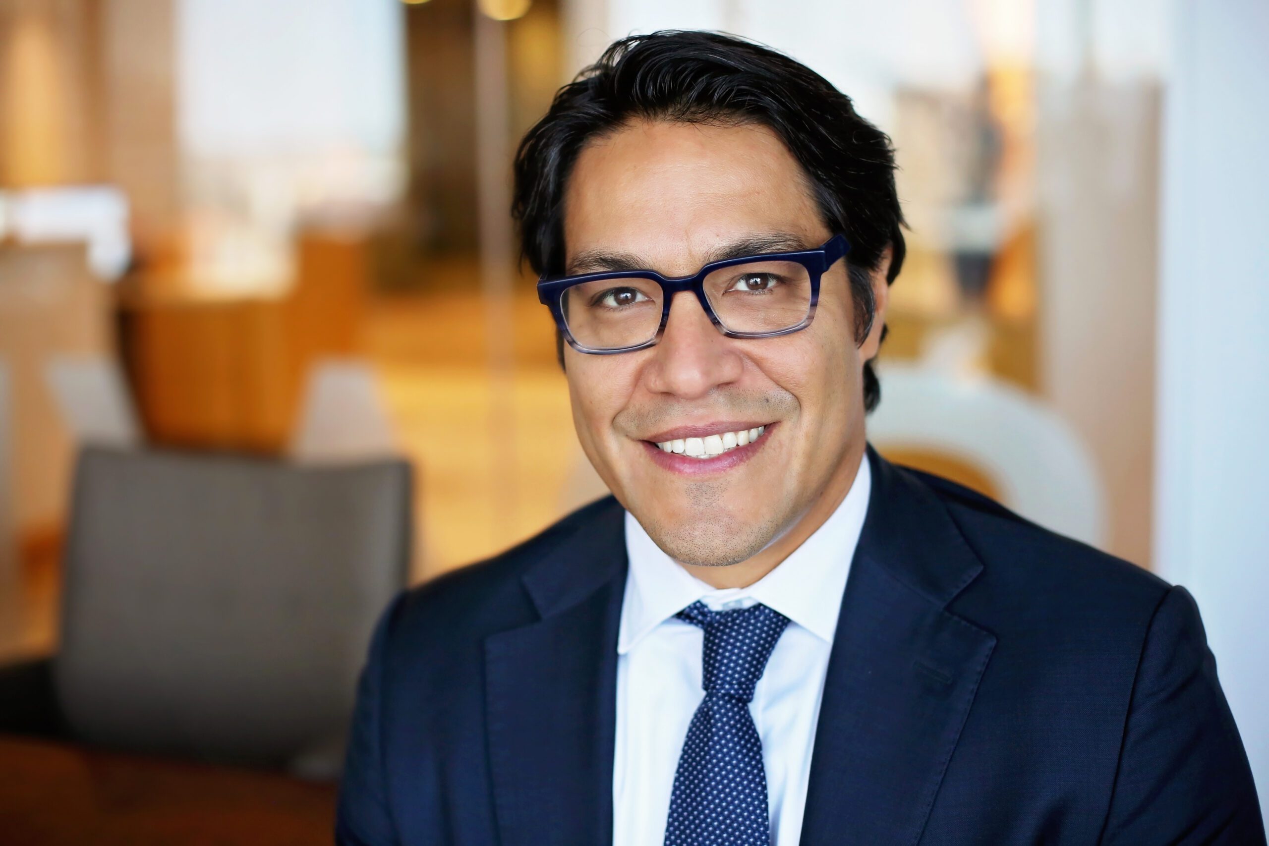 Man with black hair and glasses wearing a dark blue suit, white shirt, and blue patterned tie, smiling in an office setting with blurred background.