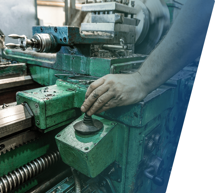A close-up of a person's hand operating a lever on an old, green industrial lathe machine. The machine shows signs of wear and rust, with various metal components and adjustment knobs visible. The background is slightly blurred, emphasizing the hand and the lever.