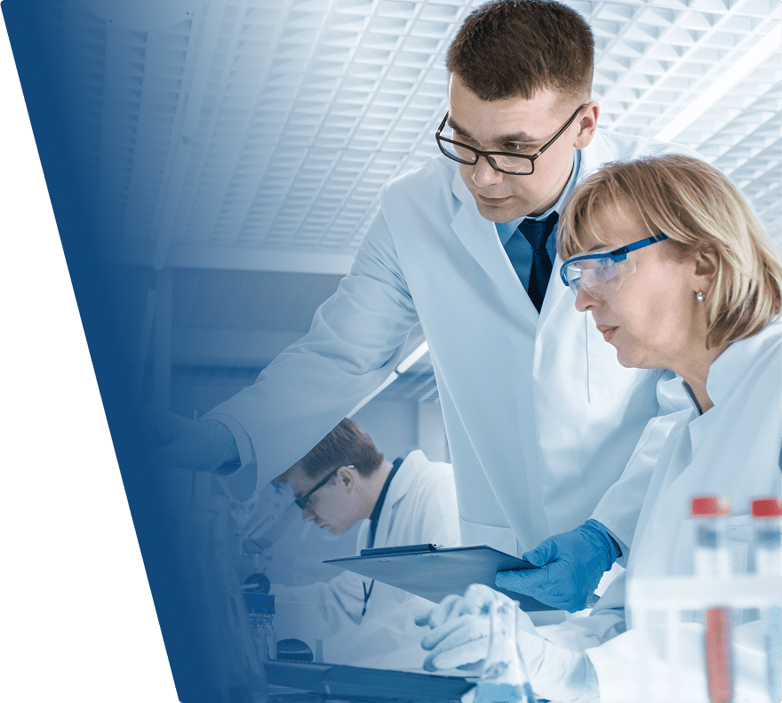 Two scientists in white lab coats and protective eyewear are working together in a laboratory. One is holding a clipboard and pointing at something, while the other is focused on the task. Laboratory equipment and test tubes are visible in the foreground. Another scientist is working in the background.