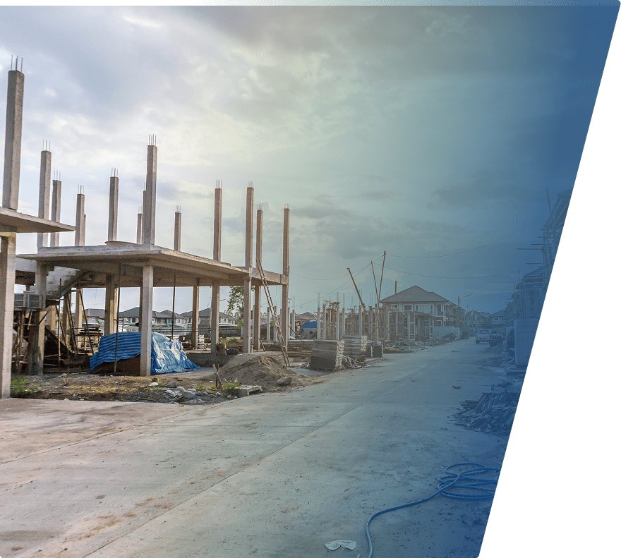 Partially constructed concrete buildings with exposed vertical columns and scaffolding along a paved road, under a cloudy sky. Construction materials and equipment are scattered around the site.