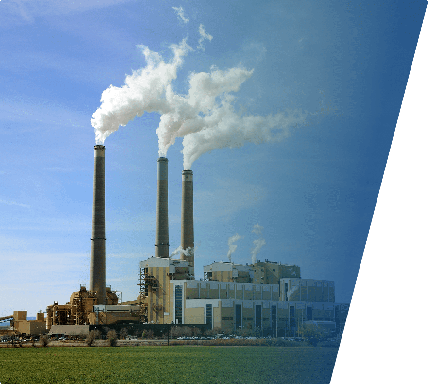 A large industrial power plant with three tall smokestacks emitting thick white smoke against a clear blue sky. The facility consists of multiple beige and gray buildings with various structures and equipment. In the foreground, there is a grassy area.