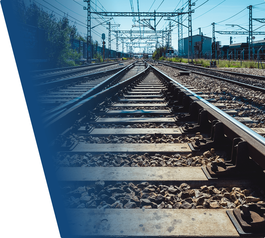 Railway tracks converging in the distance under a clear blue sky, with overhead electrical lines and poles running parallel to the tracks. Gravel and concrete sleepers support the rails, and industrial buildings and fencing are visible on the right side. Trees and greenery line the left side of the tracks.