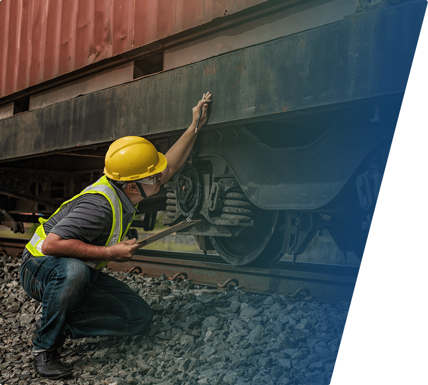 A worker wearing a yellow hard hat, safety goggles, and a reflective vest is crouched down inspecting the underside of a train car. The worker holds a clipboard in one hand and is pointing or touching the train car's metal frame with the other hand. The train is on railway tracks surrounded by gravel.