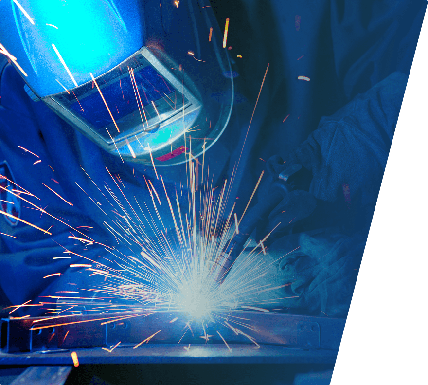 A person wearing a blue welding helmet and protective gloves is welding metal, producing bright sparks and intense light at the welding point. The background is dark, emphasizing the bright welding activity.